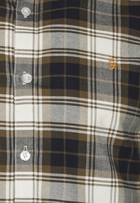 Koszula w kratę w odcieniach brązu, zieleni i czerni, wykonana z tkaniny o gładkiej fakturze. Pojedyncze haftowane logo w kolorze pomarańczowym.
