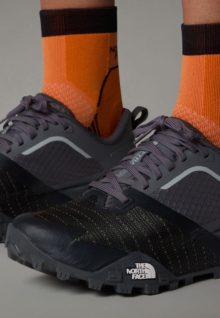 Chaussures de course grises et noires avec une tige en mesh, des détails texturés et une semelle épaisse. Portées avec des chaussettes couper-court orange présentant un motif.