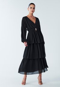 Schwarzes mehrlagiges Kleid mit tiefem V-Ausschnitt, langen Ärmeln und einem elastischen Taillenband mit goldenem Schnallendetail. Transparenter Stoff mit fließender Silhouette.