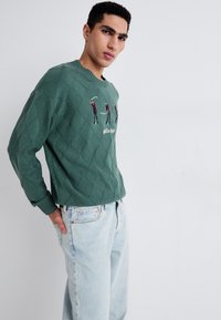 Groene gebreide trui met een diamanten patroon en geborduurde golfspelers. Gepaard met lichtblauwe denim jeans. Casual fit.