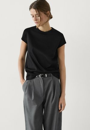 Donna con top a maniche corte nero infilato in pantaloni grigi a vita alta con pieghe e cintura nera, in piedi contro uno sfondo bianco semplice.