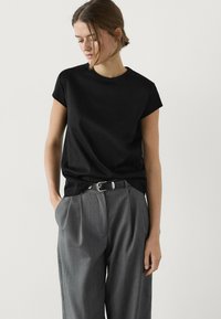 Žena ve černém topu s krátkým rukávem, který má zastrčený do šedých vyplétaných kalhot s vysokým pasem a černým páskem, stojící před jednobarevným bílým pozadím.