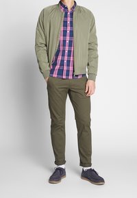 Olivgrön bomberjacka, rutig skjorta i rosa och blå nyanser, slim olivfärgade byxor, grå mockaskor, vita strumpor, modern avslappnad outfit.