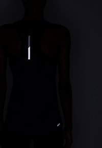 Person som bär en mörk ärmlös idrottstop med en reflexvertikal remsa på övre ryggen och en reflexlogotyp nära den nedre sidan.