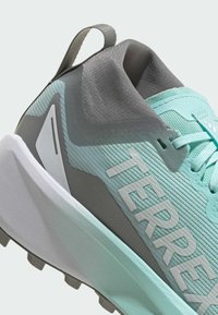 Jasnoniebieski sportowy but z teksturowaną siateczkową cholewką, szarymi akcentami, pętelką do zakładania oraz białymi napisami, wyposażony w amortyzowaną podeszwę i wypustki zapewniające przyczepność.