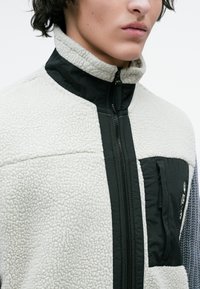 Close-up de uma pessoa a usar uma jaqueta de sherpa branca com um bolso de peito com zip preto e um colarinho alto.