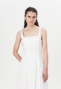 Robe en lin blanc avec un décolleté carré et de larges bretelles, dotée d'un corsage ajusté et d'une jupe fluide, texture lisse et design minimal.