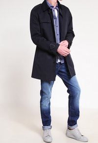 Svart trenchcoat med krage, knappstängning och sidofickor, bärs över en blåmönstrad skjorta och blå jeans. Vita sneakers.