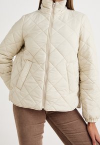 Beige quiltet jakke med høj krave, lange ærmer og lynlås foran. Har to sidelommer og en blød, glat tekstur.