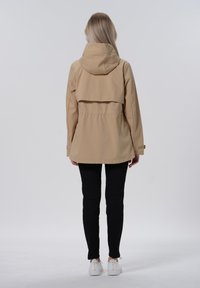 Beige wasserdichte Jacke mit Kapuze, taillierter Passform und Rückenschlitzdetail. Kombiniert mit schwarzen Hosen und weißen Sneakers.