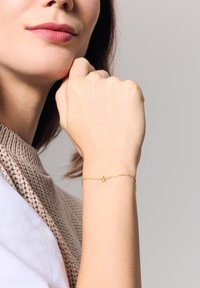 Złota bransoletka łańcuszkowa z małym charms'em w kształcie litery "A", noszona na dłoni na neutralnym tle. Bransoletka ma delikatną, subtelną fakturę.