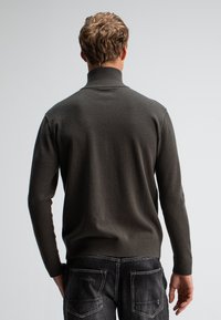 Dunkelgrüner Strickpullover mit hohem Kragen, gerippten Bündchen und Saum. Getragen mit dunklen Jeans, von hinten fotografiert.