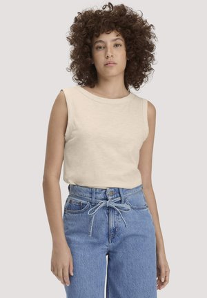 Frau mit lockigem Haar, die ein ärmelloses beiges Oberteil und eine hoch taillierte blaue Jeans mit einer Schleife aus Denim trägt, stehend vor einem einfarbigen Hintergrund.