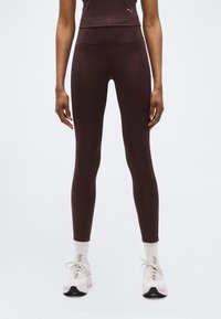 Άτομο που φοράει σφιχτό κολάν σκούρου μωβ χρώματος, άσπρες κάλτσες και λευκά αθλητικά παπούτσια, στέκεται μπροστά σε απλό φωτεινό φόντο.