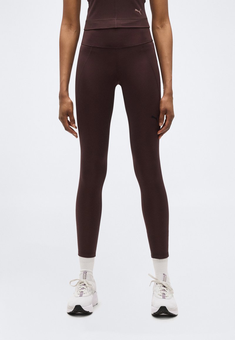 Άτομο που φοράει σφιχτό κολάν σκούρου μωβ χρώματος, άσπρες κάλτσες και λευκά αθλητικά παπούτσια, στέκεται μπροστά σε απλό φωτεινό φόντο.