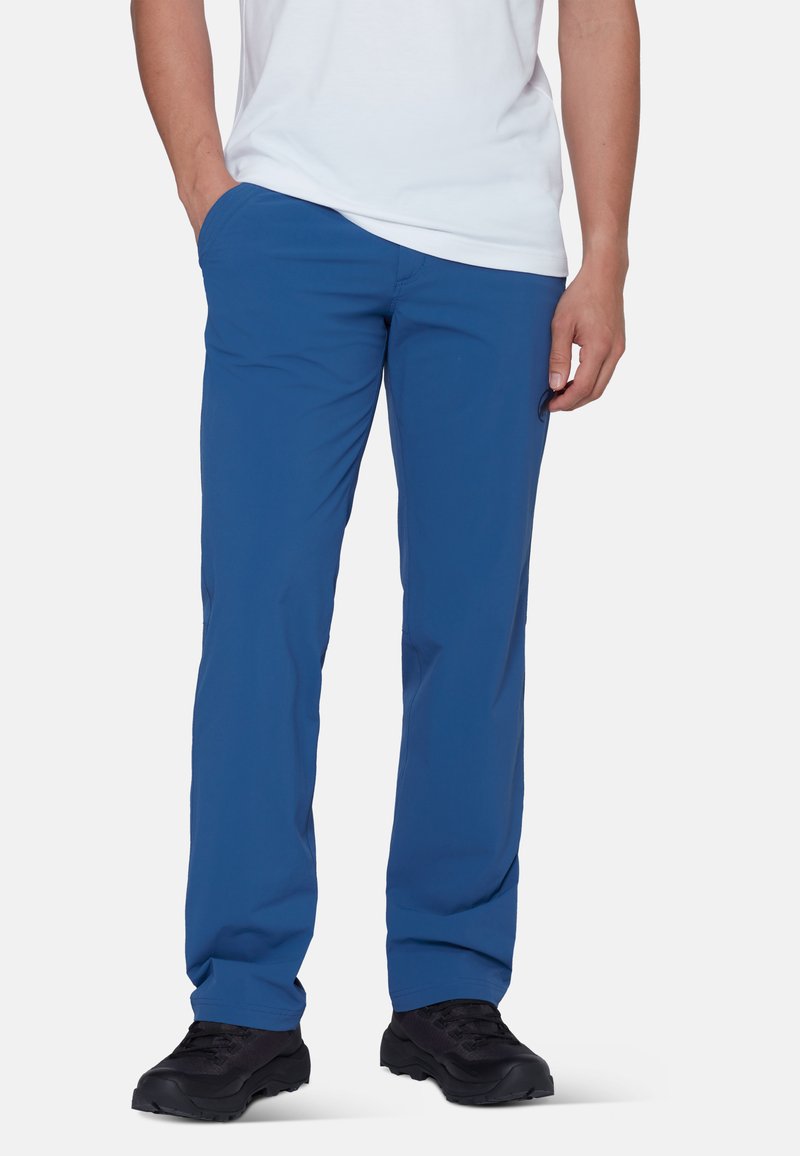 Homme portant un pantalon droit bleu, des chaussures de randonnée noires et une chemise blanche à manches courtes, debout avec une main dans la poche.