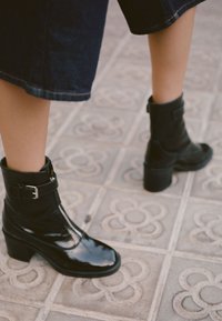 Czarne lakierowane botki za kostkę z kwadratowym noskiem, sznurówką z przodu oraz regulowanym zapięciem. Gruba obcas oraz teksturowana podeszwa.