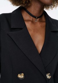 Zwarte dubbel-breasted blazer met een gladde textuur en knoopsgaten. Voorzien van glanzende knopen en een zwarte choker met een hart hanger.