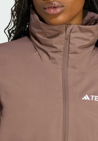 Veste zippée marron avec col montant et tissu texturé. Équipée d'une fermeture éclair métallique et d'un petit logo blanc sur la poitrine.
