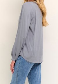 Blouse gris clair à manches longues avec une texture lisse. Elle présente un ourlet arrondi et des poignets boutonnés. Assortie à un jean bleu taille haute.