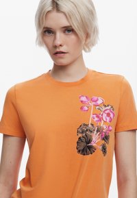 T-shirt arancione a maniche corte in cotone, caratterizzato da fiori e foglie ricamati in rosa sul lato sinistro, texture liscia, colletto a girocollo.