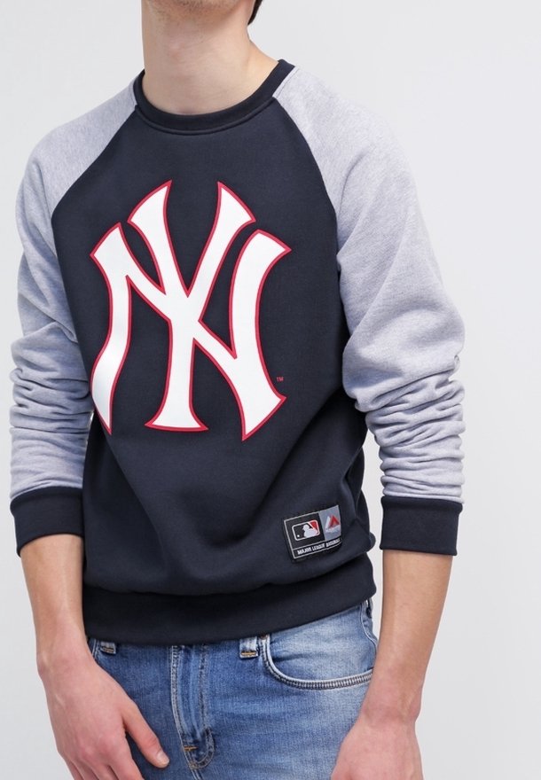 Personne portant un sweat-shirt noir et gris avec un grand logo stylisé "NY" blanc et rouge, et un jean bleu.