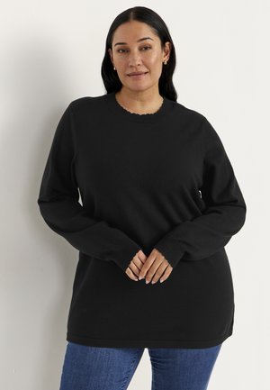 Kvinde med langt mørkt hår iført en sort langærmet sweater og blå jeans stående mod en ensfarvet lys baggrund.