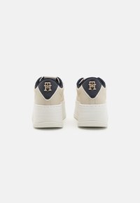 Beżowe sneakersy z granatowymi akcentami, skórzana cholewka o teksturowanej powierzchni, gładka biała podeszwa oraz złote logo na pięcie. Widok z tyłu, bez dodatkowych szczegółów.