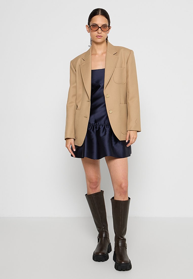 Beige op maat gemaakte blazer, marineblauw satijnen mini-jurk met plooien, en kniehoge bruine chunky haklaarzen. Transparante zonnebril als accent.