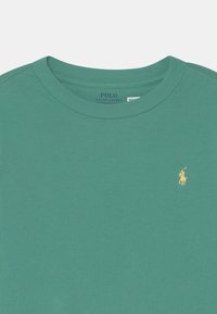 Grünes Baumwoll-T-Shirt mit Rundhalsausschnitt, das ein kleines gelbes gesticktes Logo auf der linken Brust zeigt. Weiche Textur, lässige Passform.