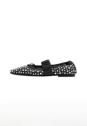 Zapato plano negro con punta afilada, adornado con tachuelas plateadas, presentando una correa de moño de satén en la parte superior. Textura suave y perfil bajo.