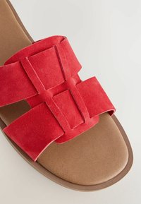 Røde ruskinds slide-sandaler med brede stropper krydset i midten, hvilende på en brunt læderlignende sål med en glat tekstur.