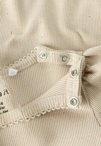 Tessuto a coste beige con glitter argento, caratterizzato da un bordo scallop e bottoni a pressione. Etichetta di cura attaccata, che mostra taglia e istruzioni per il lavaggio.