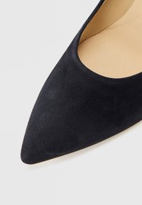 Svart semsket skinnpumps med spiss tupp og åpne sider, med en glatt beige indre fôring og solid hæl.