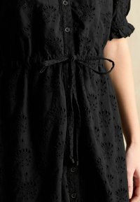 Das schwarze, bestickte Kleid verfügt über florale Muster, kurze Ärmel, eine Knopfleiste und einen Kordelzug in der Taille. Der strukturierte Stoff verleiht Tiefe.