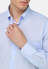 Jasnoniebieska koszula w kratę zapinana na guziki, z szerokim kołnierzem, długimi rękawami i kieszenią na piersi. Materiał wydaje się gładki i lekki.