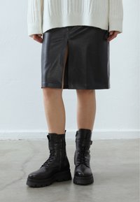 Jupe en cuir noir avec une fente frontale, associée à des bottines en dentelle noires avec une semelle robuste et des sangles réglables. Un pull en tricot blanc par-dessus.
