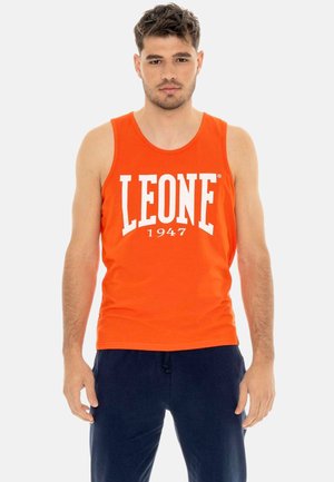 Giovane uomo con una maglietta arancione senza maniche con la scritta "LEONE 1947" e pantaloni blu scuro, in piedi davanti a uno sfondo chiaro e uniforme.