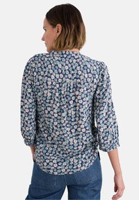 Blouse florale bleu marine avec motif de marguerites blanches, rouges et bleues. Présente des manches trois quarts et un détail de pli au dos. Texture de tissu douce.