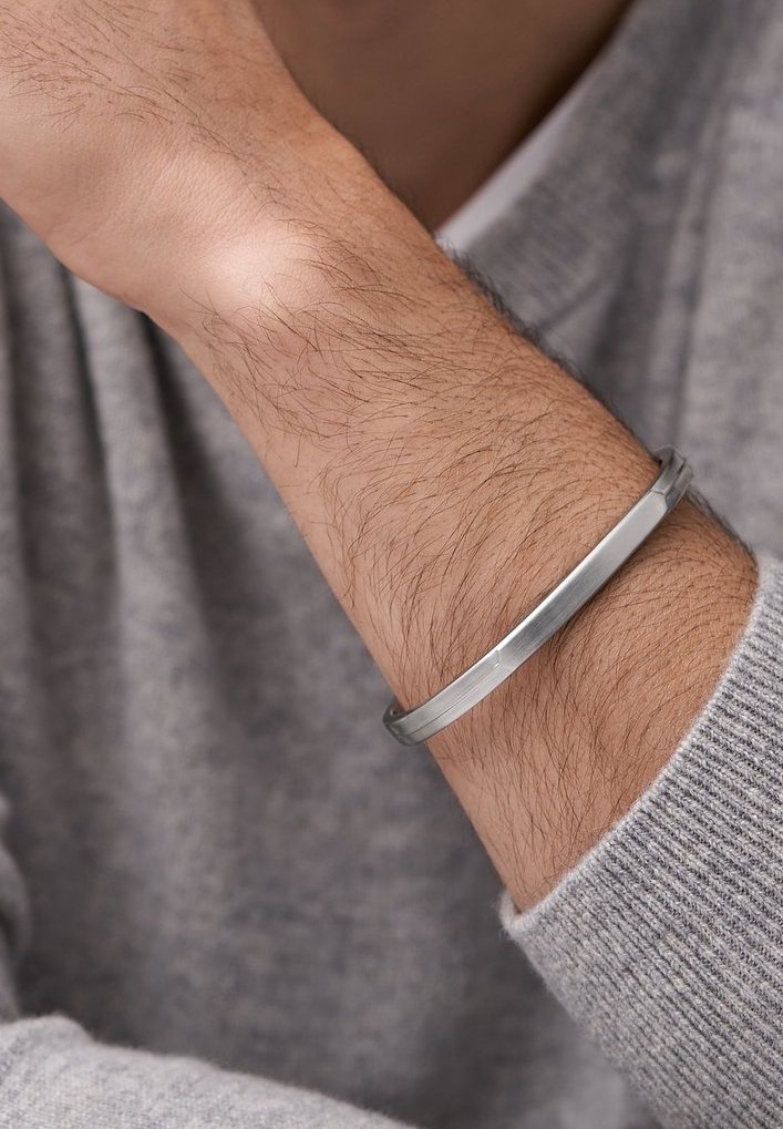 Bracelet manchette en métal argenté porté au poignet, présentant une surface lisse et plate avec un bord légèrement arrondi, sur un fond de pull gris.