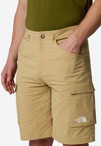 Pantaloni cargo beige in tessuto strutturato, dotati di numerose tasche e del logo The North Face ricamato in bianco sul lato inferiore.