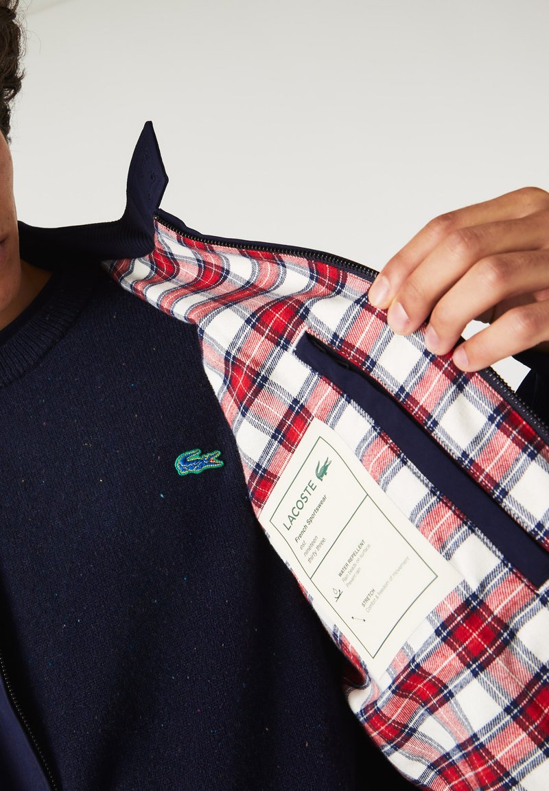 Sweatshirt marine avec des éclats multicolores, arborant un logo Lacoste vert. L'intérieur est en plaid rouge et blanc avec une étiquette d'entretien.