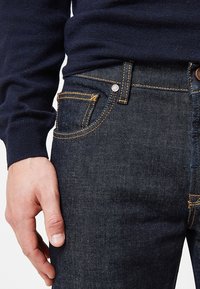 Jeans de mezclilla azul oscuro con costuras amarillas, un bolsillo frontal y un detalle de botón metálico. Combinados con un suéter azul marino, mostrando la colocación de las manos.