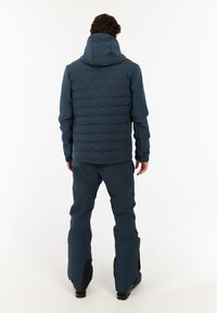 Man in een donkerblauwe gewatteerde jas met capuchon en bijpassende broek, staand met zijn gezicht van de camera af tegen een effen witte achtergrond.
