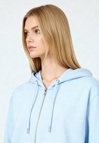 Felpa zip blu chiaro con una texture morbida, con lacci e dettagli in metallo argentato. Presenta una tasca a marsupio e una vestibilità comoda.