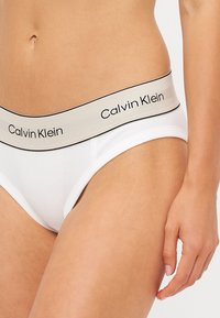Witte katoenen slips met een brede beige elastische tailleband waarop het "Calvin Klein" logo in het zwart is afgebeeld. De stof is glad met een lichte stretch.