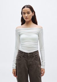Top met lange mouwen en off-shoulder ontwerp in licht wit stof met geplooide textuur, gecombineerd met een high-waist, donkerbruine broek met muntenzakje.