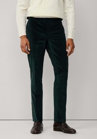 Muž v tmavozelených kordových nohaviciach, smotanovom svetrí s copánkovým vzorom a tmavohnedých kožených mokasínach, stojaci proti jednofarebnému svetlému pozadiu.