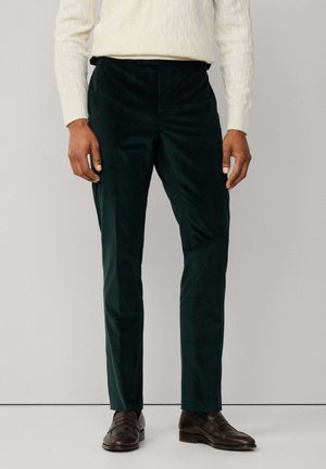 Mann in dunkelgrünen Cordhosen, cremefarbigem Strickpullover und dunkelbraunen Lederloafers, der vor einem schlichten, hellen Hintergrund steht.