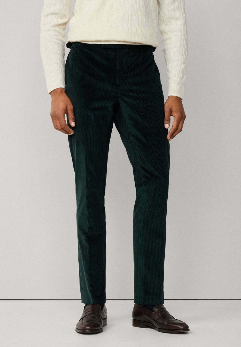 Muž v tmavozelených kordových nohaviciach, smotanovom svetrí s copánkovým vzorom a tmavohnedých kožených mokasínach, stojaci proti jednofarebnému svetlému pozadiu.